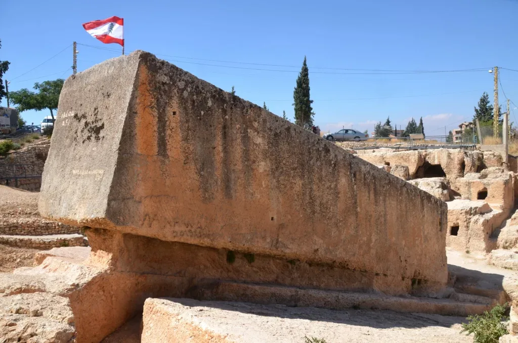 Ogroman antički kameni megalit, poznat kao 'Kamen trudne žene', koso postavljen u pješčanom kamenolomu u Baalbeku, sa libanskom zastavom na vrhu pod vedrim plavim nebom.