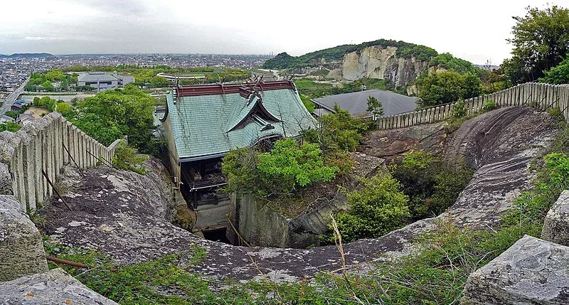 Pogled odozgo na istorijski lokalitet kamenoloma, koji prikazuje tradicionalnu japansku građevinu sa prepoznatljivim plavo-zelenim krovom. U daljini se prostire moderni grad i strmi zidovi kamenoloma prekriveni zelenilom.